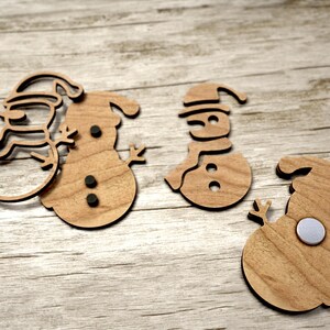 May include: Three wooden snowman decorations with black felt accents. The decorations are cut out in the shape of a snowman with a hat and scarf. The decorations are on a light brown wooden surface.