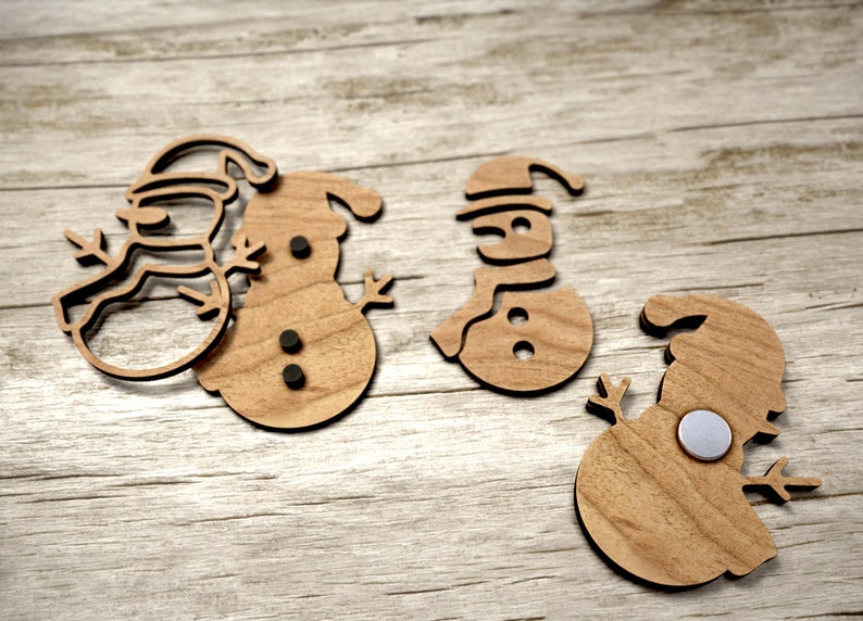 May include: Three wooden snowman decorations with black felt accents. The decorations are cut out in the shape of a snowman with a hat and scarf. The decorations are on a light brown wooden surface.