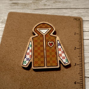 May include: A wooden cutout of a brown hooded jacket with a red heart on the front and colourful patterned sleeves. The jacket is on a brown background.
