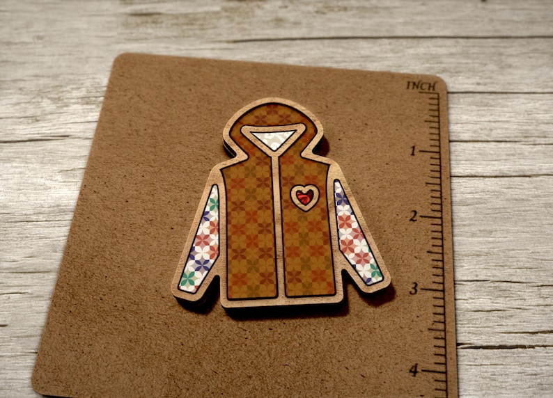 May include: A wooden cutout of a brown hooded jacket with a red heart on the front and colourful patterned sleeves. The jacket is on a brown background.