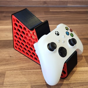 May include: A white Xbox controller with black buttons and a red and black honeycomb-patterned stand.