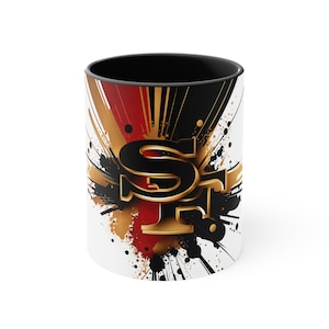 May include: White ceramic mug with a black rim and handle. The mug features a graphic design of the San Francisco 49ers logo in gold and black with red and black paint splatters.