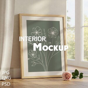 Könnte beinhalten: Ein Holzrahmen mit grünem Hintergrund und einer weißen Linienzeichnung von Blumen. Der Text "INTERIOR MOCKUP" ist auf dem Rahmen gedruckt. Der Rahmen steht auf einem Holzboden mit einer rosa Rose.