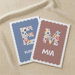 Könnte beinhalten: Zwei personalisierte Karten mit gewellten Rändern. Eine Karte ist blau mit einem floral gemusterten Buchstaben "E" und dem Namen "Elea" in Weiß. Die andere Karte ist mauvefarben mit einem floralen "M" und dem Namen "Mia".