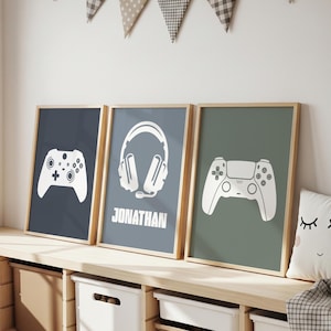 Könnte beinhalten: Drei gerahmte Drucke mit Gaming-Motiven. Jeder Druck zeigt eine weiße Grafik auf farbigem Hintergrund: ein Controller auf Marineblau, Kopfhörer mit dem Namen "JONATHAN" auf Blau-Grau und ein Controller auf Olivgrün. Die Drucke werden auf einem Holzregal ausgestellt.