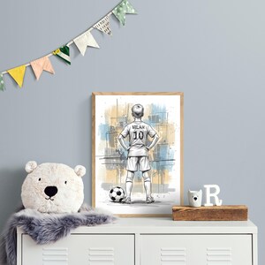 May include: A framed art print featuring a sketch of a soccer player in a Milan jersey with the number 10. A soccer ball is at the player's feet. The artwork is displayed on a white cabinet with a plush bear, a wooden block, and decorative flags.