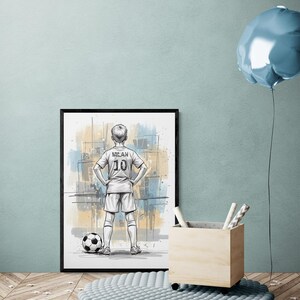 May include: Framed artwork featuring a soccer player in a white jersey with "MILAN 10" on the back, standing with hands on hips next to a soccer ball. The background has abstract blue and yellow paint strokes. A soccer-themed art print.