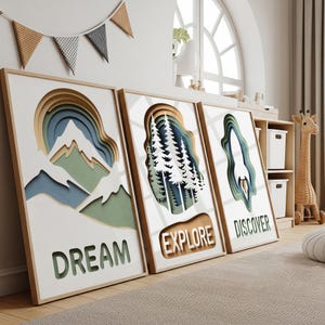 Könnte beinhalten: Drei gerahmte Wandkunstwerke mit Naturmotiven. Jedes Stück zeigt ein mehrschichtiges Design mit ausgeschnittenen Formen von Bergen, Bäumen und einer Rakete. Die Wörter "DREAM", "EXPLORE" und "DISCOVER" sind auf jedem Rahmen zu sehen.