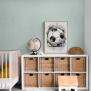 May include: A framed art print of a soccer ball in a child's room. The artwork is in black, white, and gold tones, with abstract background elements. The room features a white storage unit with woven baskets, a globe, and a small wooden house.