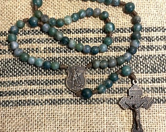 May include: A green agate rosary with a bronze St. Michael medal and crucifix. The rosary is laid out on a brown burlap background.