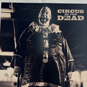 Puede incluir: Una fotografía en blanco y negro de un payaso sosteniendo un martillo grande. El payaso lleva un traje de payaso tradicional con una gran sonrisa. La foto está firmada "Bill Moseley" y "CIRCUS OF THE DEAD".