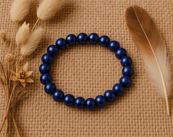 Può includere: Un braccialetto di perline blu navy. Il braccialetto è composto da perline rotonde e lucide. Il braccialetto è esposto su una superficie strutturata color beige, con fiori secchi e una piuma sullo sfondo.