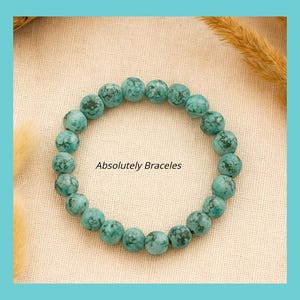 May include: A turquoise beaded bracelet with a mottled pattern. The bracelet is circular and sits on a beige surface. The text "Absolutely Bracelets" is displayed below the bracelet.