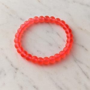 May include: A vibrant coral-colored beaded bracelet. The bracelet is made of small, round beads strung together to form a circular shape. The beads have a matte finish, giving them a soft appearance. The bracelet is displayed on a white and gray marble surface.