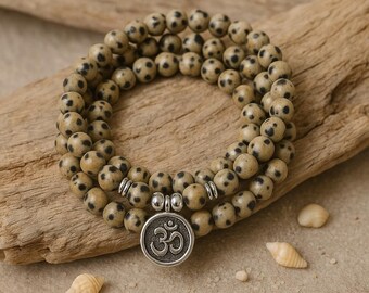 May include: A beaded necklace with a silver pendant featuring the Om symbol. The beads are beige with black spots. The necklace is resting on a piece of driftwood, with small seashells scattered around.