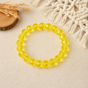Puede incluir: Un brazalete de cuentas amarillo brillante. El brazalete está hecho de cuentas facetadas y translúcidas. Las cuentas están ensartadas para formar una forma circular. El brazalete se muestra sobre una superficie de color neutro.