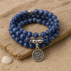 May include: A blue beaded bracelet with a silver charm featuring the Om symbol. The bracelet is made of round, dark blue beads and is wrapped around itself. The charm is circular and has a detailed design. The bracelet is displayed on a piece of driftwood.