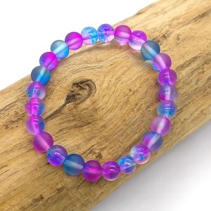 May include: A bracelet made of iridescent glass beads in shades of blue, purple, and pink. The round beads are strung together, creating a colourful accessory. The bracelet is displayed on a piece of driftwood.