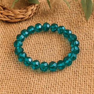 May include: A teal beaded bracelet with faceted beads. The bracelet is made of round, translucent beads strung together on an elastic band. The beads have a faceted cut, giving them a sparkling appearance. The bracelet is displayed on a textured, tan surface.