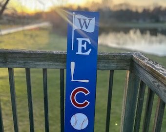 May include: A blue wooden welcome sign with white letters spelling "WELCOME". The sign features a baseball, a baseball bat, and a flag.