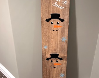 May include: A wooden sign with a brown finish features two snowman faces with black hats and orange noses. The words "Let It Snow" are written in white script. Light blue snowflakes are also painted on the sign.