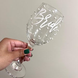 May include: A clear glass wine glass with white polka dots and the word "Bride" written in white script.