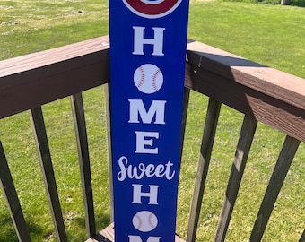 May include: A blue wooden sign with white lettering and baseball graphics. The sign reads "C", "HOME Sweet HOME" with baseballs. The sign is leaning on a wooden deck with a lake in the background.
