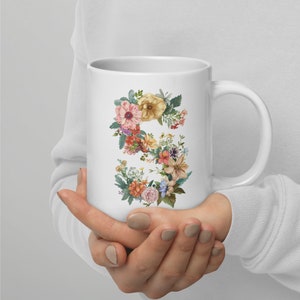 May include: White ceramic mug featuring the letter "S" formed by a colorful floral arrangement. The flowers include pink, yellow, orange, and purple blooms with green foliage. The mug has a curved handle and a smooth, glossy finish.