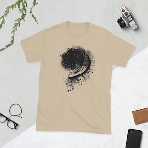 以下が含まれることがあります： ベージュのTシャツ。黒と白のピアノの鍵盤が螺旋状に描かれたグラフィック。デザインには音符と暗くテクスチャ加工された背景が含まれています。シャツは半袖で、クルーネックです。