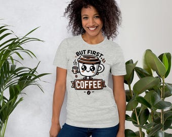 May include: A light gray t-shirt featuring a cartoon coffee cup with the text "BUT FIRST, COFFEE". The design includes a coffee bean and a small coffee cup. The shirt is worn by a person.