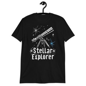 Könnte beinhalten: Schwarzes T-Shirt mit einer weißen Grafik eines Teleskops und Sternen, mit dem Text "Stellar Explorer". Das Design enthält blaue Akzente. Das T-Shirt hängt an einem schwarzen Kleiderbügel.