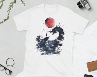 May include: A white short-sleeve t-shirt featuring a graphic print of a dark blue sea dragon rising from ocean waves towards a large red sun. A small silhouette of a person stands on a rock formation in the centre, surrounded by small birds.
