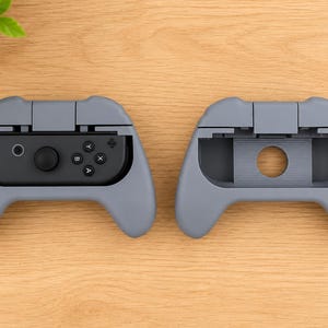 Op de afbeelding: Twee grijze gamecontroller-schelpen, &eacute;&eacute;n met een zwarte controller erin en de andere leeg. De controller heeft knoppen met de labels A, B, X en Y, en een joystick. De schelpen liggen op een houten oppervlak.