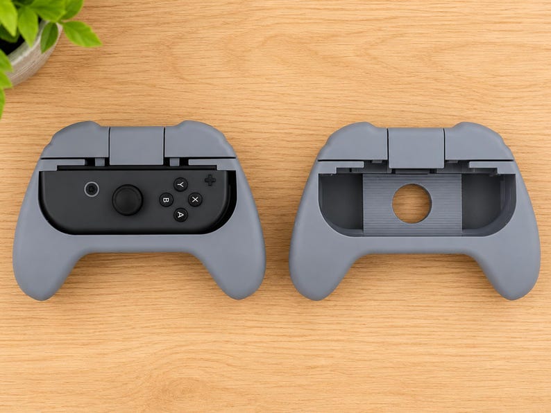 Op de afbeelding: Twee grijze gamecontroller-schelpen, &eacute;&eacute;n met een zwarte controller erin en de andere leeg. De controller heeft knoppen met de labels A, B, X en Y, en een joystick. De schelpen liggen op een houten oppervlak.
