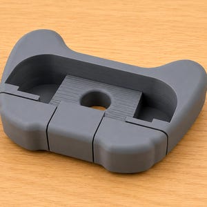 Op de afbeelding: Een grijs, 3D-geprint object met een unieke vorm, met een centrale cirkelvormige opening en een reeks rechthoekige uitsparingen. Het ontwerp lijkt een houder of een ondersteunende structuur te zijn. Het object ligt op een lichtbruin houten oppervlak.