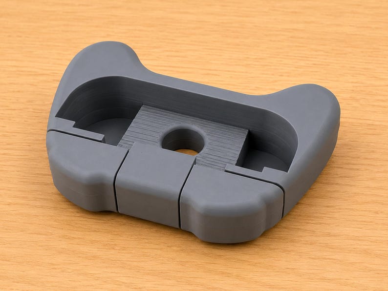 Op de afbeelding: Een grijs, 3D-geprint object met een unieke vorm, met een centrale cirkelvormige opening en een reeks rechthoekige uitsparingen. Het ontwerp lijkt een houder of een ondersteunende structuur te zijn. Het object ligt op een lichtbruin houten oppervlak.