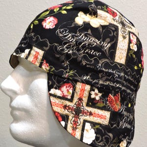 May include: Black welding cap with a floral and cross pattern. The cap features red, white, and pink flowers, butterflies, and the words "Amazing Grace" in a script font. The cap is designed to protect the wearer's head and ears.