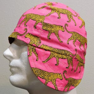 May include: A pink welding cap featuring a repeating pattern of yellow leopard print. The cap has a rounded crown and a brim that extends to cover the ears. The cap is designed to protect the head and ears from sparks and heat.