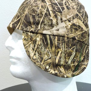 May include: A camouflage welding cap with a brown and green pattern. The cap is made of fabric and features a curved brim and ear flaps. White stitching accents the seams.
