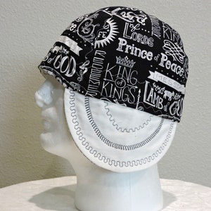 May include: Black and white patterned welding cap with the words "King of Kings" and "Prince of Peace" printed on it. The cap has a white, curved brim with black stitching and is displayed on a white mannequin head.