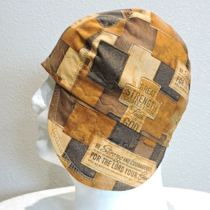 May include: A patchwork welding cap in shades of brown, tan, and black, featuring rectangular patches. The cap has text that reads "GREAT STRENGTH COMES FROM FAITH IN GOD" and "BE STRONG AND COURAGEOUS FOR THE LORD YOUR GOD."