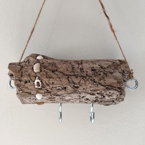 May include: A rustic, hanging key holder made from a piece of driftwood. The brown wood is adorned with small seashells and features three metal hooks for keys. It hangs from a natural rope, adding a coastal, beachy vibe.