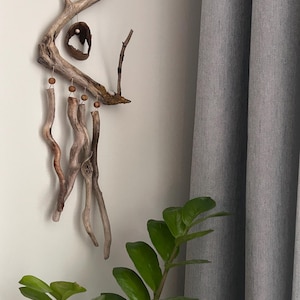 May include: A handcrafted driftwood mobile with a natural, organic aesthetic. The mobile features several pieces of driftwood, wooden beads, and hanging elements. The neutral color palette and natural materials create a calming, bohemian vibe. The mobile hangs against a white wall.