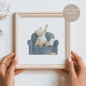 May include: A light-colored wooden frame containing a cross stitch pattern of a light brown teddy bear wearing a white striped shirt, sitting on a dark blue armchair with one arm raised, with a small stack of books beside it and text reading Cross Stitch Pattern PDF.