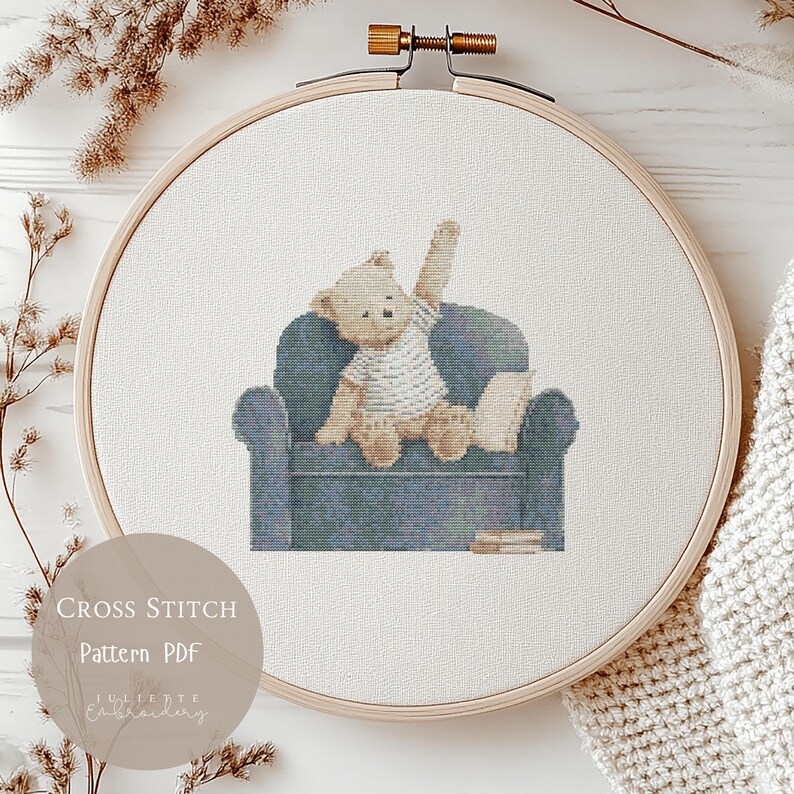 May include: A cross stitch pattern in a wooden embroidery hoop featuring a light brown teddy bear wearing a white striped shirt, sitting on a blue armchair with one arm raised, and a small stack of books beside the chair on a white fabric background.