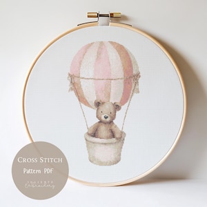 May include: Cross-stitch pattern featuring a teddy bear in a hot air balloon with pink and tan stripes. The design is set in a wooden embroidery hoop. The text "Cross Stitch Pattern PDF" is visible.