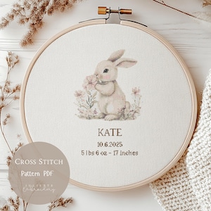 May include: A cross-stitch embroidery hoop with a light beige bunny holding flowers. The text "KATE" is below the bunny, along with the date, weight (2.5 kg), and height (43 cm). The words "Cross Stitch Pattern PDF" are on a circular label.