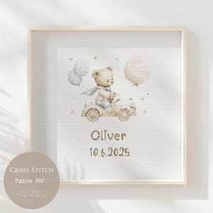 May include: Cross-stitch pattern featuring a teddy bear driving a vintage car, with balloons and stars. The name "Oliver" and the date "10.6.2025" are stitched below the car. The design is framed in a light wood frame.