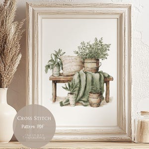 May include: Cross stitch pattern PDF for a botanical illustration of potted plants on a wooden table with a green blanket. The plants are in various sizes and shapes, with some in terracotta pots and others in wooden buckets. The illustration is in a white frame.