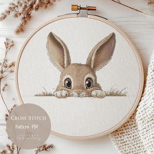 Può includere: Un modello di punto croce in PDF con un adorabile coniglietto marrone che spunta da dietro uno sfondo bianco. Il modello è intitolato "Cross Stitch Pattern PDF".